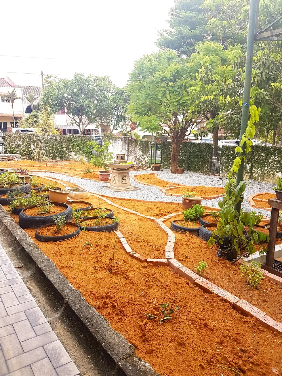 Garden with orange soil pathways, brick borders, planted tires, gravel beds, and a small stone lantern, surrounded by trees